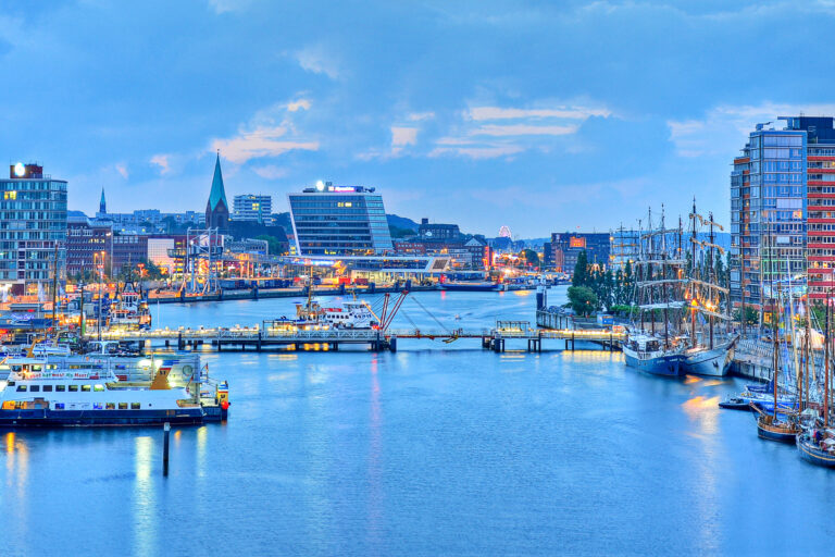 Harbor skyline at dusk with moored boats, glass buildings, and a distant Ferris wheel.