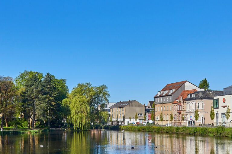 Row of buildings along a riverfront with trees on the left and a clear blue sky above, reflections on the water.