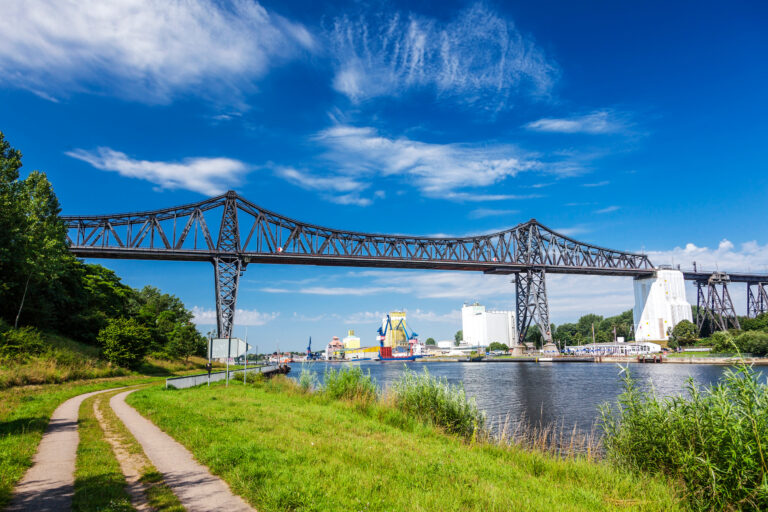 Large steel truss bridge spanning a calm river, with a grassy riverside path in the foreground and a bright blue sky above.