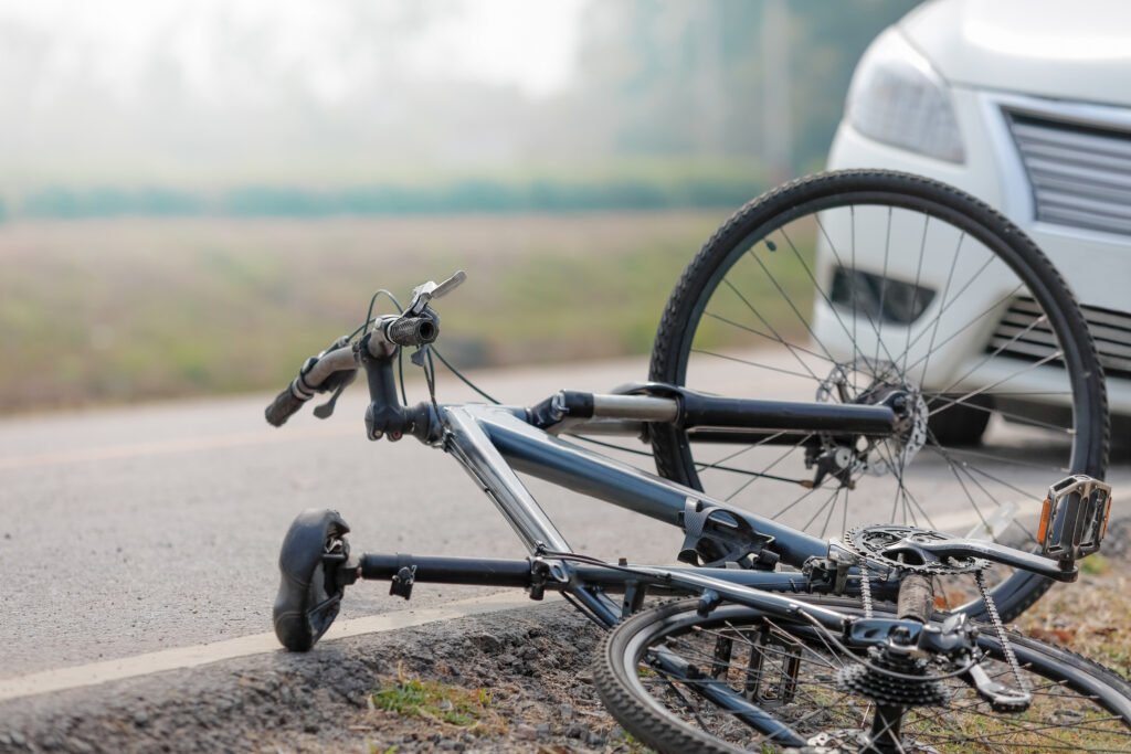 Bicycle lying on its side on the road after a crash, with a white car in the background and exposed gears visible.