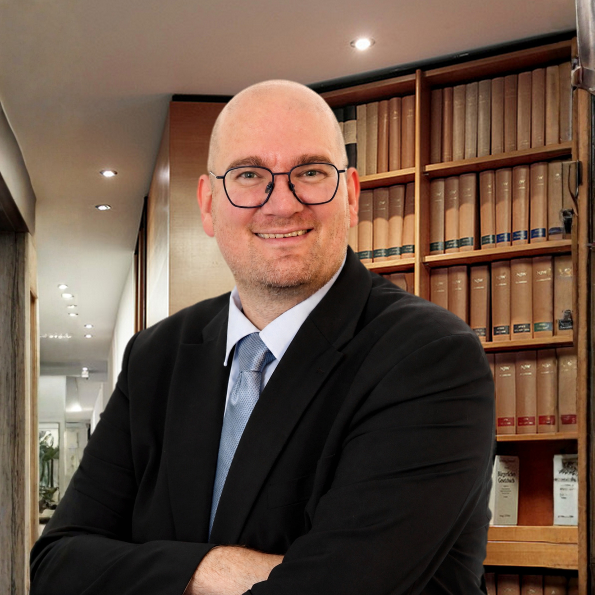Professional portrait of a smiling, bald man with glasses in a dark suit and light blue tie, standing in a library with wooden shelves of books behind him.